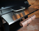 A Pimp My Grill Towel Bar for 18″ Rolls or Smaller is attached to the side shelf of a grill, holding a space-saving roll of paper.