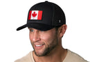 A smiling man wears a Haka Hat Canada Flag Trucker Hat—Black Canadian Snapback—with a Canadian flag patch, trimmed beard, and light shirt, standing against a plain white background.