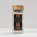 A glass jar with a wooden lid, labeled “Spice Wheel Caramel Sugar” by the brand Spice Wheel and filled with light brown caramel sugar (Net Wt. 109g), sits on a white surface against a plain background.