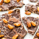 Pieces of chocolate bark topped with whole roasted almonds, swirls of milk and dark chocolate, and sprinkled with SALTOPIA Infused Sea Salts’ Saltopia Caramel Sugar: DATE NIGHT 16oz on a white surface.