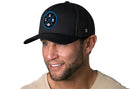 A man with light facial hair smiles while wearing the Haka Hat Carolina Trucker Hat, a black CAR X Football snapback by Haka Hat, paired with a light tan shirt. The background is plain white.