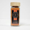 A glass jar of Spice Wheel Cayenne Pepper by Spice Wheel, featuring vibrant red cayenne pepper powder made from select chili peppers and topped with a wooden lid, rests on a white surface. The net weight is 62 grams.