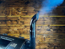 A chrome Pimp My Grill Chrome Pellet Grill Smoke Stack Trucker Series Fits GMG / Yoder Smokers attached to a grill, emitting a plume of smoke against a wood-paneled wall.