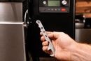 A hand holds a metal Rack Puller™ by Pimp My Grill® tool labeled HOT with a hook at one end, positioned near the control panel of a grill.