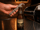 A hand holds a Rack Puller™ by Pimp My Grill® with a built-in bottle opener over a beer bottle on a stainless steel surface.