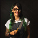 A smiling woman with long green hair, glasses, and tattoos wears a white shirt and dark apron, holding the Messermeister Chef Claudette Zepeda Chef's Knife - 7" against a dark background.