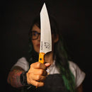 A person with tattoos and dark hair holds the Messermeister Chef Claudette Zepeda Chef's Knife - 7", featuring a yellow handle and a fish illustration on the blade, pointed toward the camera against a dark background.