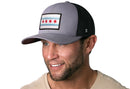 A smiling man with a short beard wears the Haka Hat Chicago Flag Trucker Hat—Gray-Black City Snapback by Haka Hat. He’s dressed in a beige shirt and faces slightly left, highlighting the prominent patch on the front of the hat.