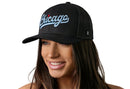 A woman with long brown hair smiles while wearing the Haka Hat Chicago Light Blue Script Trucker Hat, a black Illinois snapback featuring bold light blue script. The signature Haka Hat 5.0 design pops against a white background.