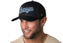 A man smiles and looks to the side, wearing a Haka Hat Chicago Light Blue Script Trucker Hat in black (Illinois snapback), with “Chicago” in light blue script on the front, against a white background.