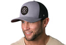A smiling man with short facial hair wears a light beige shirt and the Haka Hat Chicago Trucker Hat—a gray-black CHI X snapback mesh cap by Haka Hat, featuring an embroidered patch with crossed lines and "CHIC" on the front.