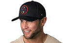 A smiling man with stubble wears the Haka Hat Chicago Trucker Hat, a black CHI X Football Snapback featuring a round logo with "CHI" and a car graphic. He pairs it with a beige crew-neck shirt against a white background.