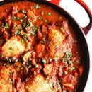 A close-up of chicken cacciatore in a red cast iron skillet, with browned chicken simmered in tomato sauce, herbs, capers, and vegetables, finished with SALTOPIA Infused Sea Salts' EUREKA SWEET PAPRIKA & Onion Salt and fresh chopped parsley.