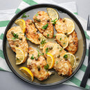 A plate of chicken piccata garnished with lemon slices, capers, and herbs is enhanced by SALTOPIA Infused Sea Salts' Lemon & Herbs Seasoning Low Sodium: TWIST AGAIN, served on a green and white striped napkin with a fork and knife beside it.