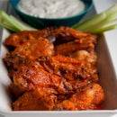A rectangular white dish holds spicy buffalo chicken wings tossed in SALTOPIA Infused Sea Salts' Saltopia Hot Honey, served with fresh celery sticks and a bowl of creamy ranch or blue cheese dip in the background.