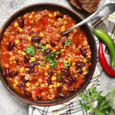 A bowl of chili with ground meat, kidney beans, corn, tomatoes, and peppers is seasoned with SALTOPIA Taco Seasoning: HOLY MOLY for authentic flavor, garnished with parsley and served with bread and whole red and green chili peppers.