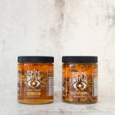 A Spice Witch Chili Crisp Box of 2 by Spice Witch is shown, featuring one Spicy Honey and one Chili Crisp jar side by side on a light surface with a neutral background.