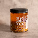 A box of 2 Spice Witch Chili Crisp jars—handcrafted chili oil with crispy flakes, sealed with black lids—sits on a light gray surface against a neutral background.