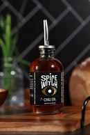 A glass bottle of Spice Witch Chili Oil by Spice Witch, topped with a metal pour spout, rests on a wooden surface. Blurred kitchen items and a tiled wall in the background evoke its savory avocado oil-infused flavor.