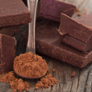 A close-up of SALTOPIA Infused Sea Salts' Saltopia Chocolate Sugar: HEYO COCOA 16oz in a metal spoon, with dark chocolate chunks on a rustic wooden background.