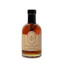 A clear glass bottle of Under The Bark Syrup Co Cinnamon Maple Syrup, filled with amber syrup and a cinnamon stick. The maple leaf label reads "100% Pure Maple Syrup, Cinnamon" and notes it's made in Minnesota.