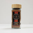 A glass jar from the brand Spice Wheel, labeled "Spice Wheel Cinnamon Sugar," with a wooden lid, containing 100 grams of a blend made from granulated sugar and ground cinnamon, is displayed against a plain white background.