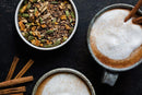 Two mugs of chai latte with frothy tops sit next to a bowl of loose World Spice Classic Chai and several cinnamon sticks on a dark surface, highlighting the rich aroma of this signature blend by World Spice, made with robust black tea and fragrant spices.