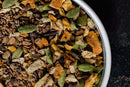 A close-up of a bowl filled with a blend of dried spices and herbs—including green cardamom pods, cloves, dried orange peel, and chopped ginger—the essential ingredients in World Spice Classic Chai from World Spice—set against a dark background.