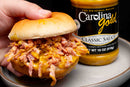 A hand holds a pulled pork sandwich topped with tangy mustard-based Gourmet Carolina Gold BBQ sauce, positioned next to a matching bottle of the Gourmet Carolina Gold Mix-and-Match 6-Pack of Gourmet Carolina Gold BBQ Sauces.