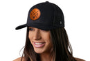 A woman with long brown hair smiles while wearing the Haka Hat Cleveland Trucker Hat Leather in black, featuring a round brown CLE X patch. The background is white.
