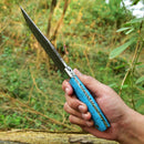 A person holds the Texan Knives Custom Handmade Damascus Skinning Knife – 4.5" Blade with a blue patterned handle outdoors among greenery, showcasing its striking Damascus steel blade—ideal for outdoor enthusiasts.