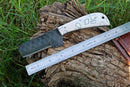 A Texan Knives Custom Handmade Cowboy Bull Cutter Knife with a 4" patterned J2 steel blade and a white geometric handle rests on a tree stump beside a metal ruler, green grass in the background.