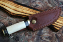 The Texan Knives Custom Handmade Damascus Camping Knife with a camel bone handle is partially sheathed in a stitched brown leather case, featuring brass details and resting on a camouflage-patterned surface.