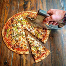 Someone slices a vegetable and meat-topped pizza on a wooden table using the Texan Knives Axe Pizza Cutter—custom hand-engraved with a J2 steel blade. One slice is separated from the rest.
