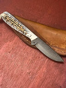The Texan Knives Handmade Buckaroo Belt Knife features an 8” rasp steel blade, an epoxy pine cone handle, and a western leather sheath, displayed on a reddish wooden surface.