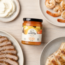A jar of Texan Heartland Coconut Pineapple Habanero Hot Jam centered among plates of sliced meat, shrimp, and cream cheese.