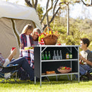 A family enjoys a picnic by their tent, sharing food on the DirectDeals Folding Camping Table with 2-Tier Open Shelves for Outdoor BBQ, surrounded by lawn chairs on the grass.