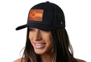 A woman with long brown hair smiles while wearing the Haka Hat Colorado Flag Trucker Hat Leather, a black CO snapback by Haka Hat, featuring a bold state flag patch. The background is plain white.
