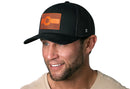 Smiling man with a short beard wears a Haka Hat Colorado Flag Trucker Hat Leather in black, featuring an orange flag patch and a light-colored shirt, posed against a white background.
