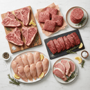 An assortment of raw meats from the Dude Food Common Man Meat Box by Dude Food—including Kansas City Strip Steaks, American Ground Beef, burger patties, sliced beef, Boneless Chicken Breasts, and pork chops—artfully arranged on plates and boards and garnished with fresh herbs and lemon wedges.