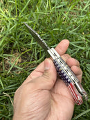 A hand holds the Texan Knives Lone Star 3-Inch Damascus Pocket Knife, custom handmade with a bone handle adorned in an American flag pattern, set against green grass.