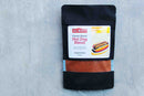 A black pouch labeled World Spice Coney Island Hot Dog Blend, featuring an illustration of a hot dog, rests on a light gray surface. Created by World Spice and inspired by the original Coney Island hot dog recipe, the pouch has a clear bottom that reveals the reddish spice blend inside.