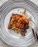 A grilled pork chop with defined grill marks rests on a white plate with grey stripes, its savory exterior brushed lightly with Tuffy Stone Original BBQ Sauce with Chipotle. A fork and knife are placed on the plate, while the meat is garnished with a few small herbs and a delicate white flower.