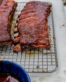 Two racks of glazed, cooked ribs rest on a metal cooling rack over a white surface. In the lower left corner, a blue bowl holds Tuffy Stone Original BBQ Sauce with Chipotle. The ribs are shiny and appear freshly sauced with Tuffy Stone’s signature barbecue sauce.