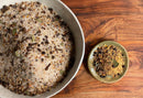 Two bowls rest on a wooden surface: the larger white bowl holds World Spice’s Corned Beef Cure & Cook Kit—a coarse seasoning blend with bay leaves, peppercorns, and spices, ideal for preparing a St. Patrick's Day meal or crafting your own homemade corned beef; the smaller green bowl contains a ground version of the same flavorful spice mix from World Spice.
