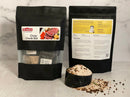 Two black pouches labeled World Spice Corned Beef Cure & Cook Kit sit on a counter; one displays the front featuring ingredients, while the other shows instructions on the back. In front of them are a scoop of spice mix and scattered spices—perfect for preparing homemade corned beef or as a convenient, easy-to-use addition to your St. Patrick’s Day meal from World Spice.
