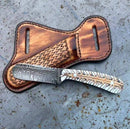 The Cowboy Bull Cutter Knife by Texan Knives features a custom handmade Damascus steel blade and feather-engraved handle, resting on a tooled leather sheath with intricate designs atop a weathered metal surface.