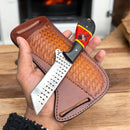 A hand holds the Texan Knives Premium Cowboy Bull Cutter Knife with an 8" rasp steel tanto blade and a red and black handle, above a brown ranch pancake sheath with decorative stitching, on a wooden surface.