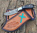 The Texan Knives Cowboy Bull Cutter features a 4.5" Damascus steel blade and sheep horn handle, resting on a brown leather sheath with blue cross and embossed oak leaf, displayed on textured tree bark.