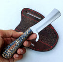 A hand holds the Texan Knives Cowboy Bull Cutter Knife – a custom handmade J2 steel blade with a patterned handle and wide, rectangular blade. A dark brown leather sheath with red stitching and engravings lies behind it.
